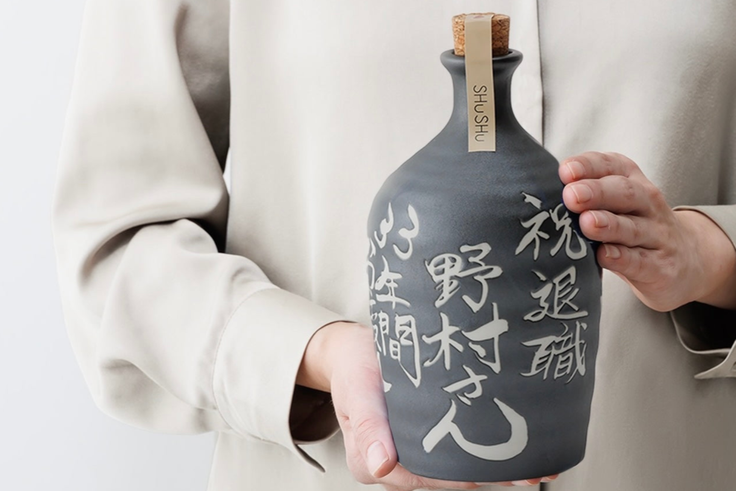 「祝 退職 野村さん」と職人が手書きした「いぶし銀」カラーの美濃焼陶器ボトルを、両手で丁寧に持つ様子。高級感のある贈り物。